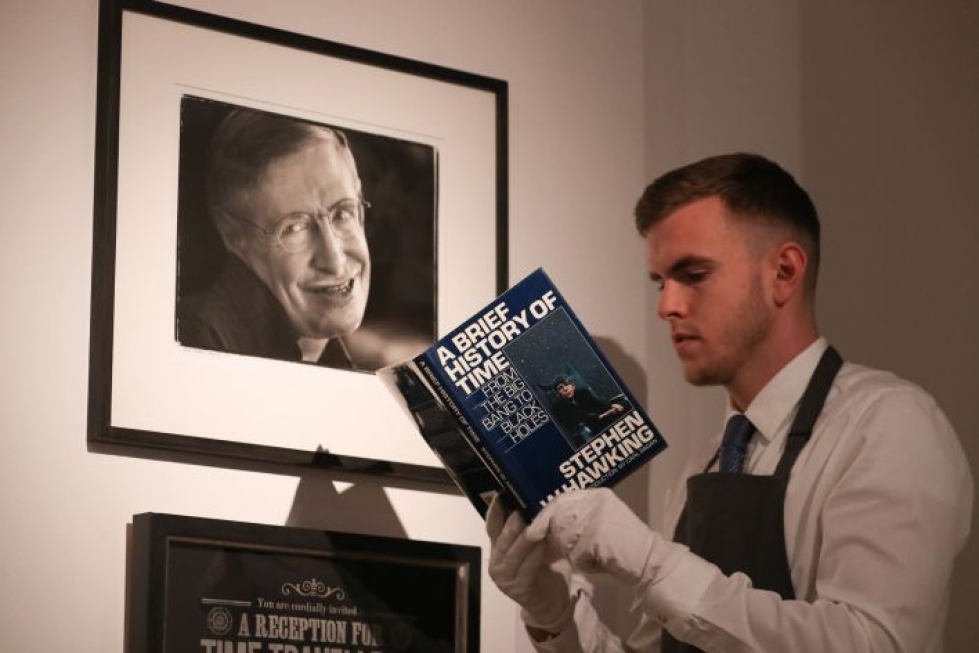 Christie'sin työntekijä käsittelee fyysikko Stephen Hawkingin (seinän valokuvassa) jäämistöön kuuluvaa Ajan lyhyt historia -teosta huutokauppakamarilla Lontoossa. LEHTIKUVA / AFP